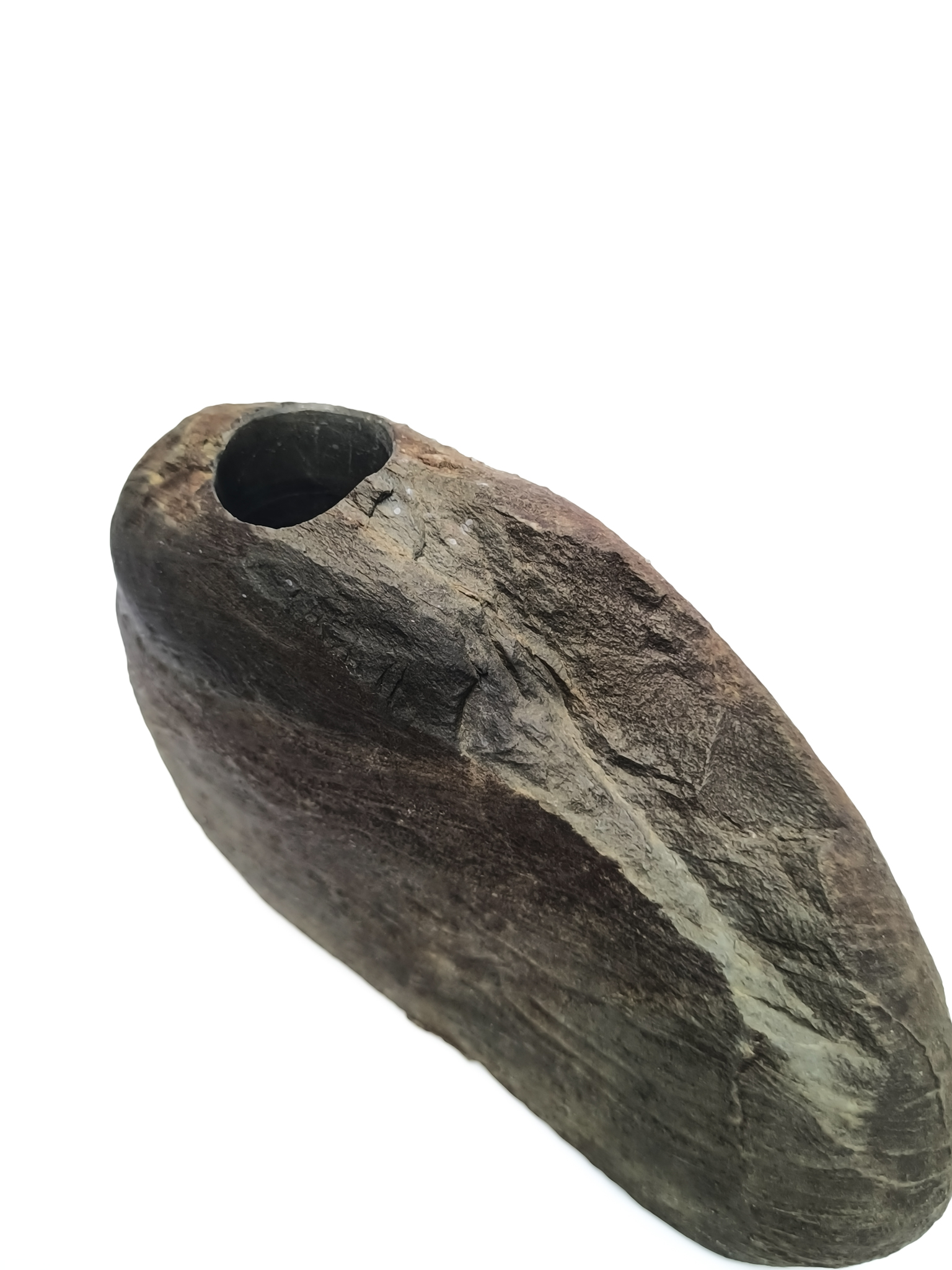 Низкая асимметричная массивная каменная ваза тёмно-серого, коричневого и чёрного цветов с шероховатой, рельефной и матовой поверхностью, украшенной горизонтальными и диагональными волнистыми слоями разной толщины, создающими рельефный узор, и цветовым переходом от тёмного, почти чёрного, оттенка в верхней части к серо-коричневому на основной поверхности.